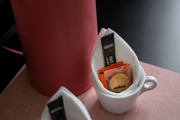 Una taza blanca que contiene un paquete de café Lavazza y una pequeña galleta, sobre una superficie rosa junto a un recipiente rojo.