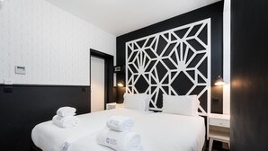 Habitación de hotel con una cama grande, ropa de cama blanca, un panel de pared geométrico, una mesita de noche y una mesita auxiliar.