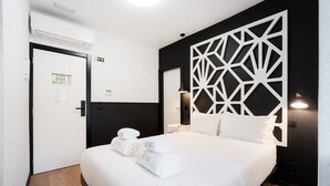 Una habitación de hotel con una cama, una lámpara geométrica fijada a la pared y una puerta con letrero de salida.