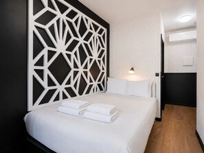 Une chambre d’hôtel dotée d’un panneau mural géométrique moderne, d’un lit soigneusement fait avec du linge de lit blanc et d’une lampe de chevet.