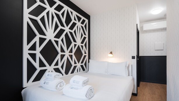 A hotel room with a bed, white towels, a geometric wall panel, a bedside lamp, and a door leading to another room.