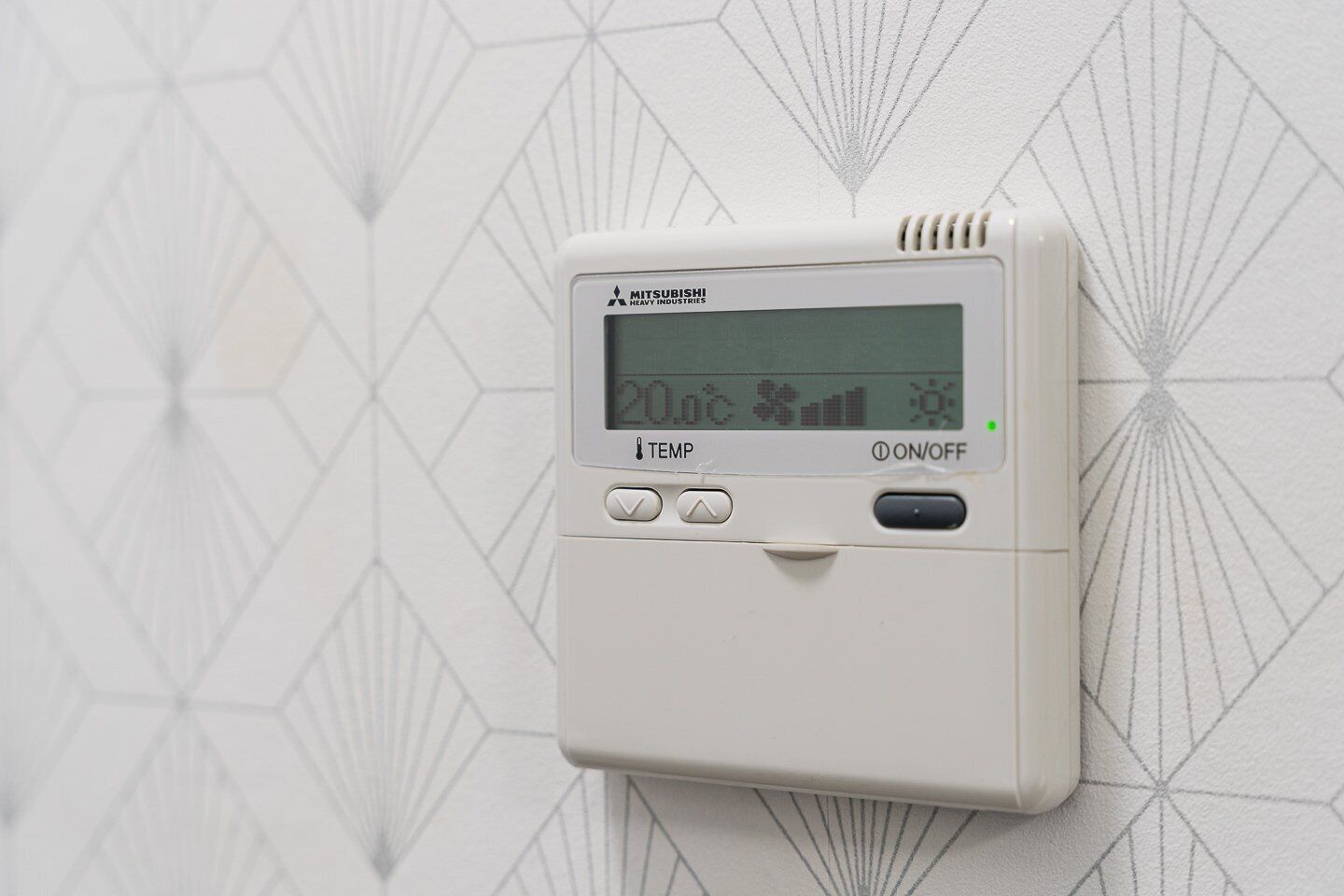 Een witte Mitsubishi Heavy Industries thermostaat bevestigd aan een muur met een geometrisch patroon.