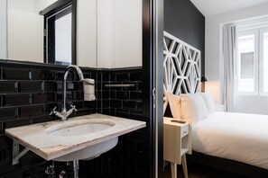 Une chambre d’hôtel moderne avec un lavabo en marbre, un mur recouvert de carreaux noirs et blancs, une tête de lit aux motifs géométriques et une grande fenêtre.