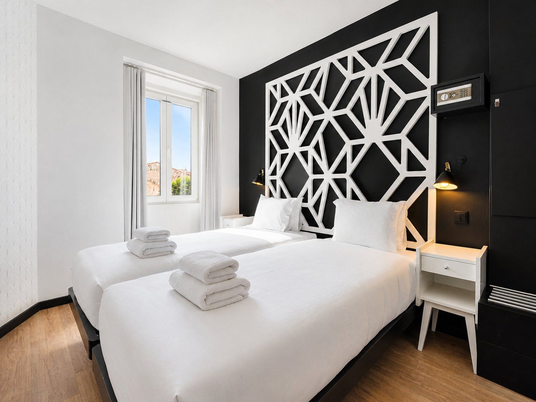 A hotel room with two beds, a large window, a geometric wall panel, and a bedside table with a lamp.