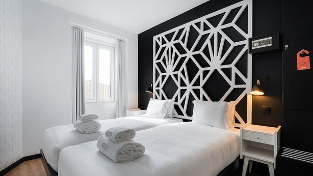 A hotel room with two beds, white linens, a geometric wall panel, a bedside table with a lamp, and a red tag indicating a relaxation menu. - The Twenties by GuestReady - Room 14 (Lisbon)