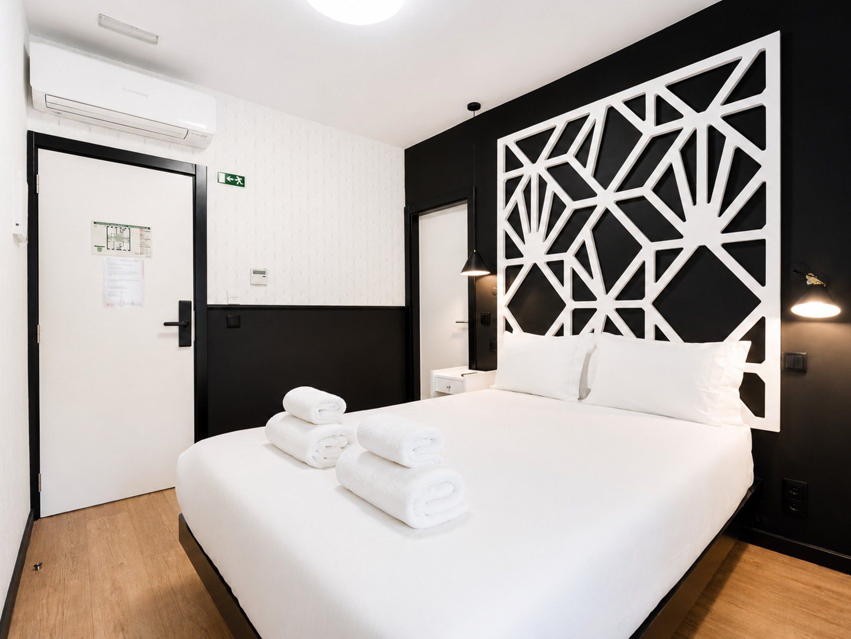 A hotel room with a bed, a wall-mounted air conditioner, a door with a sign, and a decorative geometric panel.