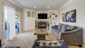 A living room with a fireplace, a sofa, a coffee table, and a television. - Deep Blue! GameRoom, Playground & Seasonal Pool (Colorado Springs)