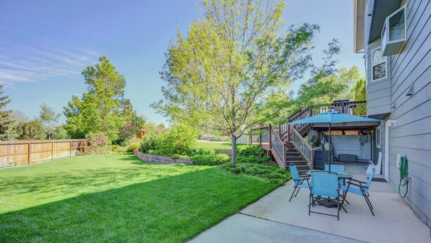 A backyard with a lawn, a tree, a patio with chairs, and a covered area with stairs leading to a deck. - Deep Blue! GameRoom, Playground & Seasonal Pool (Colorado Springs)