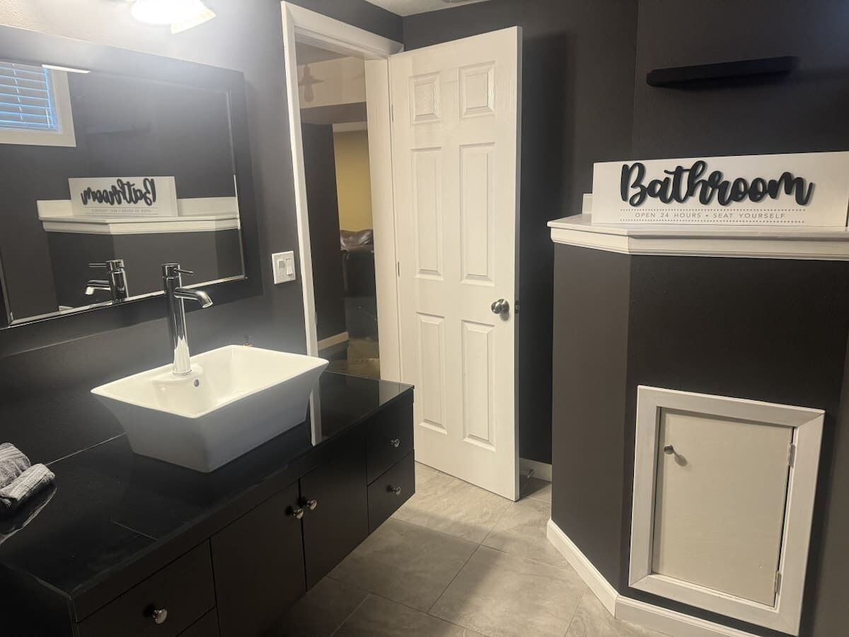 A modern bathroom with a black and white color scheme, featuring a large mirror, a white sink, and a sign indicating the bathroom is open 24 hours.