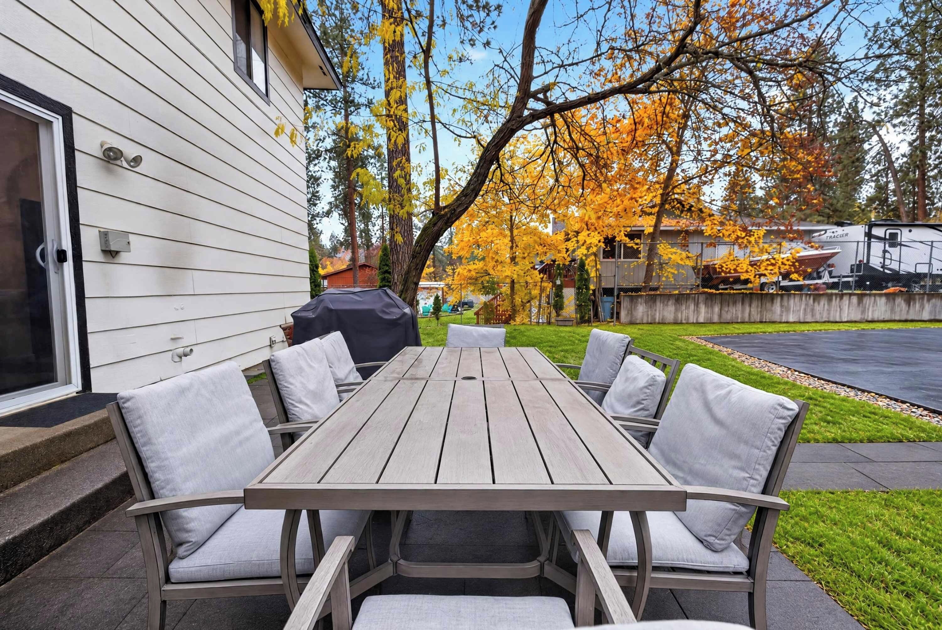 Une terrasse extérieure avec une table et des chaises en bois, un barbecue abrité et une vue sur un quartier résidentiel aux arbres aux couleurs automnales.