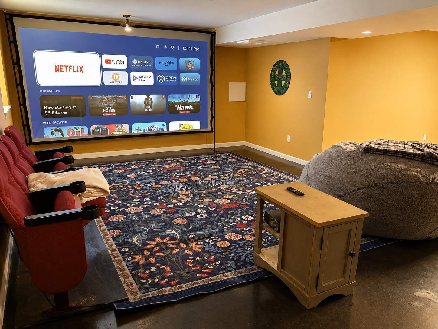 A home theater room with a large screen displaying streaming services, a red couch, a bean bag, and a small wooden table.