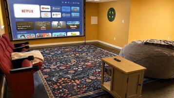 A home theater room with a large screen displaying streaming services, a red couch, a bean bag, and a small wooden table.