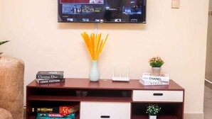 A flat-screen TV mounted on a wall displaying various streaming services. Below the TV, there's a wooden console with a vase of yellow flowers, a small potted plant, and a few books.
