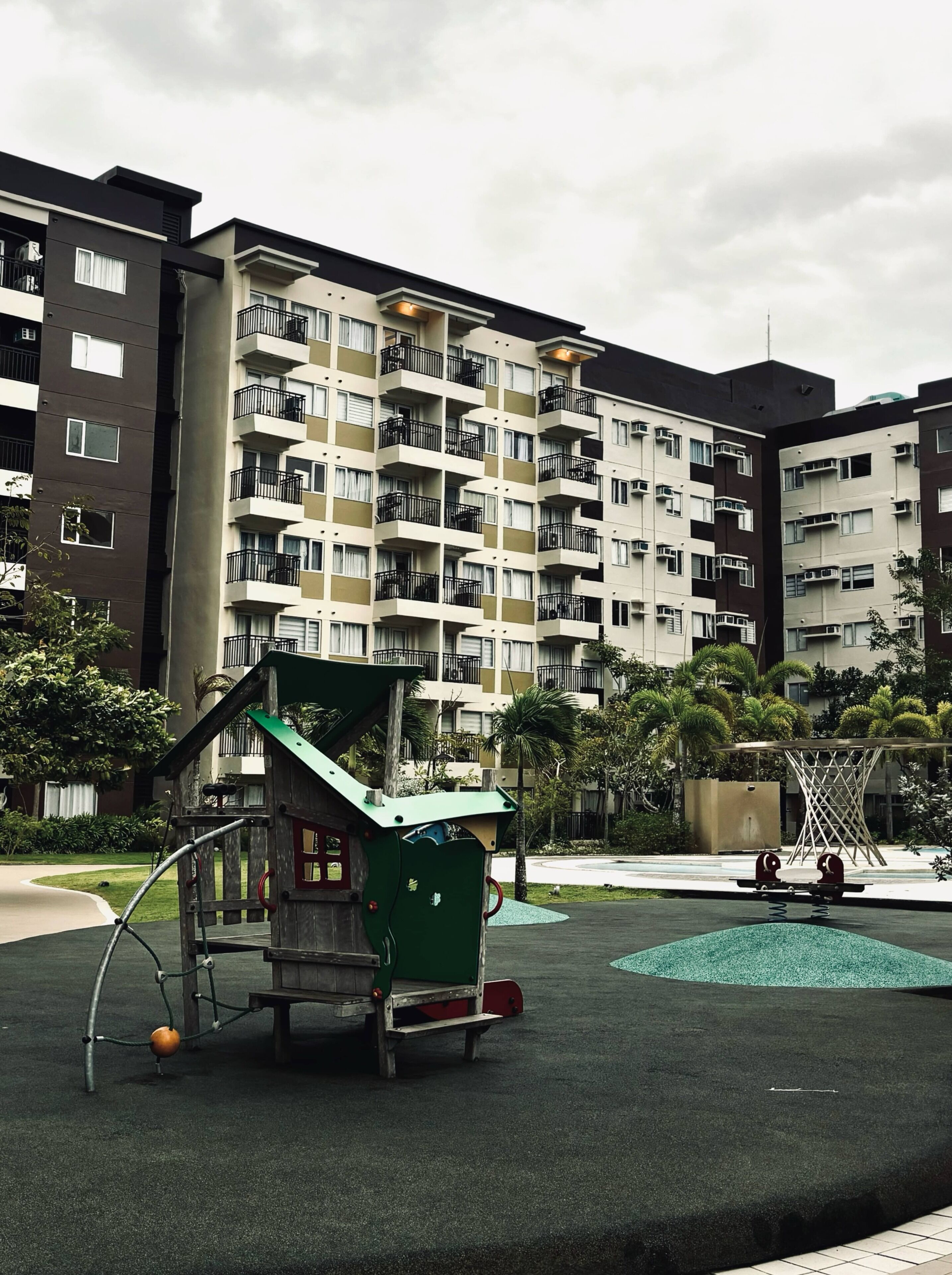 A playground with a green playhouse and a basketball court, surrounded by multi-story residential buildings.