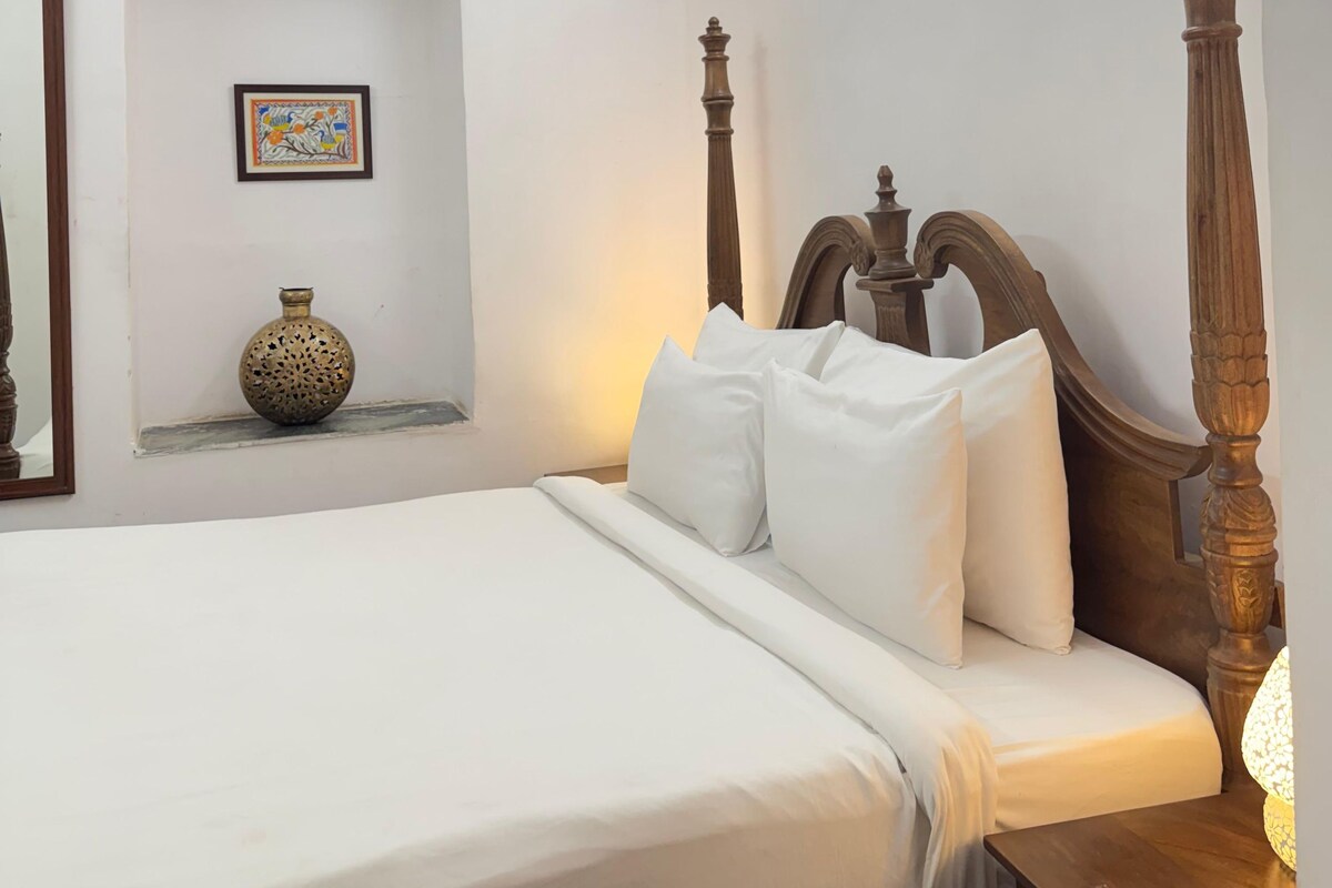 Quarto com estrutura de cama de madeira, roupa de cama branca, mesa de cabeceira com abajur, ventilador de teto e quadro pendurado na parede.