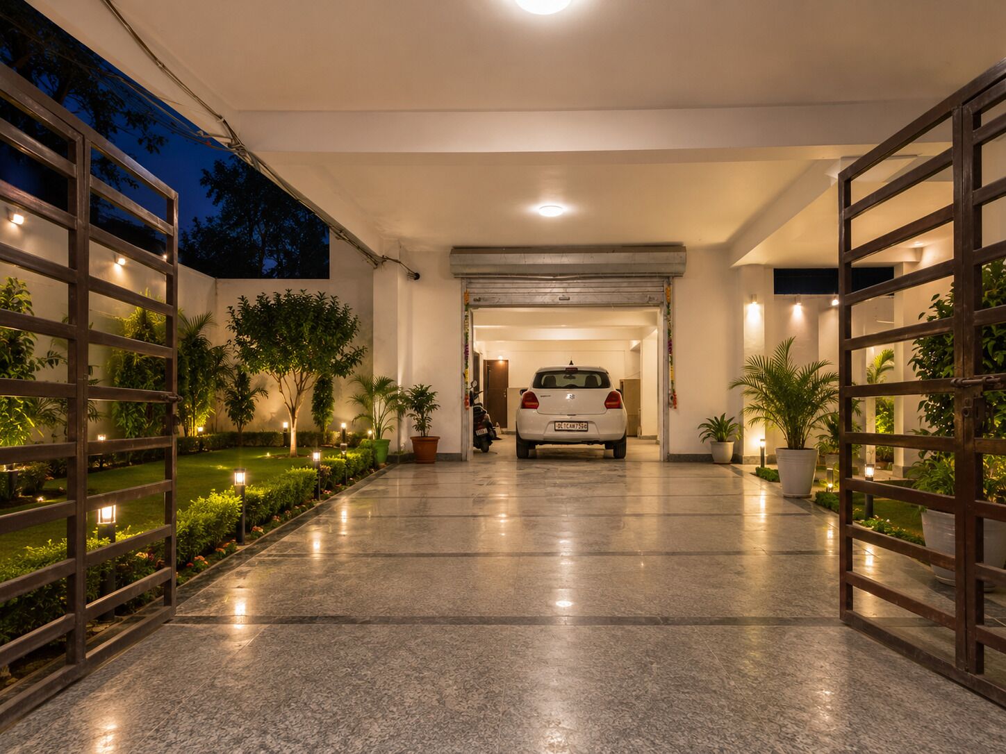 Une allée bien éclairée mène à un garage où une voiture est garée, le tout étant agrémenté de plantes en pot et doté d’un sol carrelé impeccable.