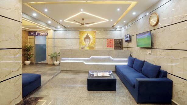 A lobby with a reception desk, a blue sofa, a coffee table, a clock, and a wall-mounted TV. - Hotel Volga Deluxe (New Delhi)