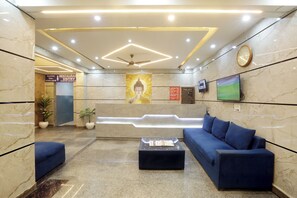 A lobby with a reception desk, a blue sofa, a coffee table, a clock, and a wall-mounted TV. - Hotel Volga Deluxe (New Delhi)