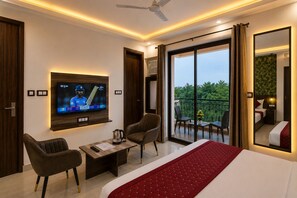 A hotel room with a television, a bed, a desk with a chair, and a balcony with a view of greenery. - Hotel Volga Deluxe (New Delhi)