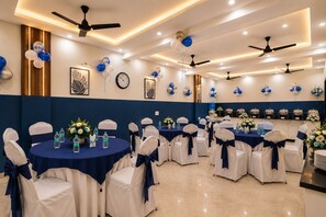 A banquet hall with round tables, chairs, ceiling fans, wall-mounted clocks, and buffet stations. - Hotel Volga Deluxe (New Delhi)