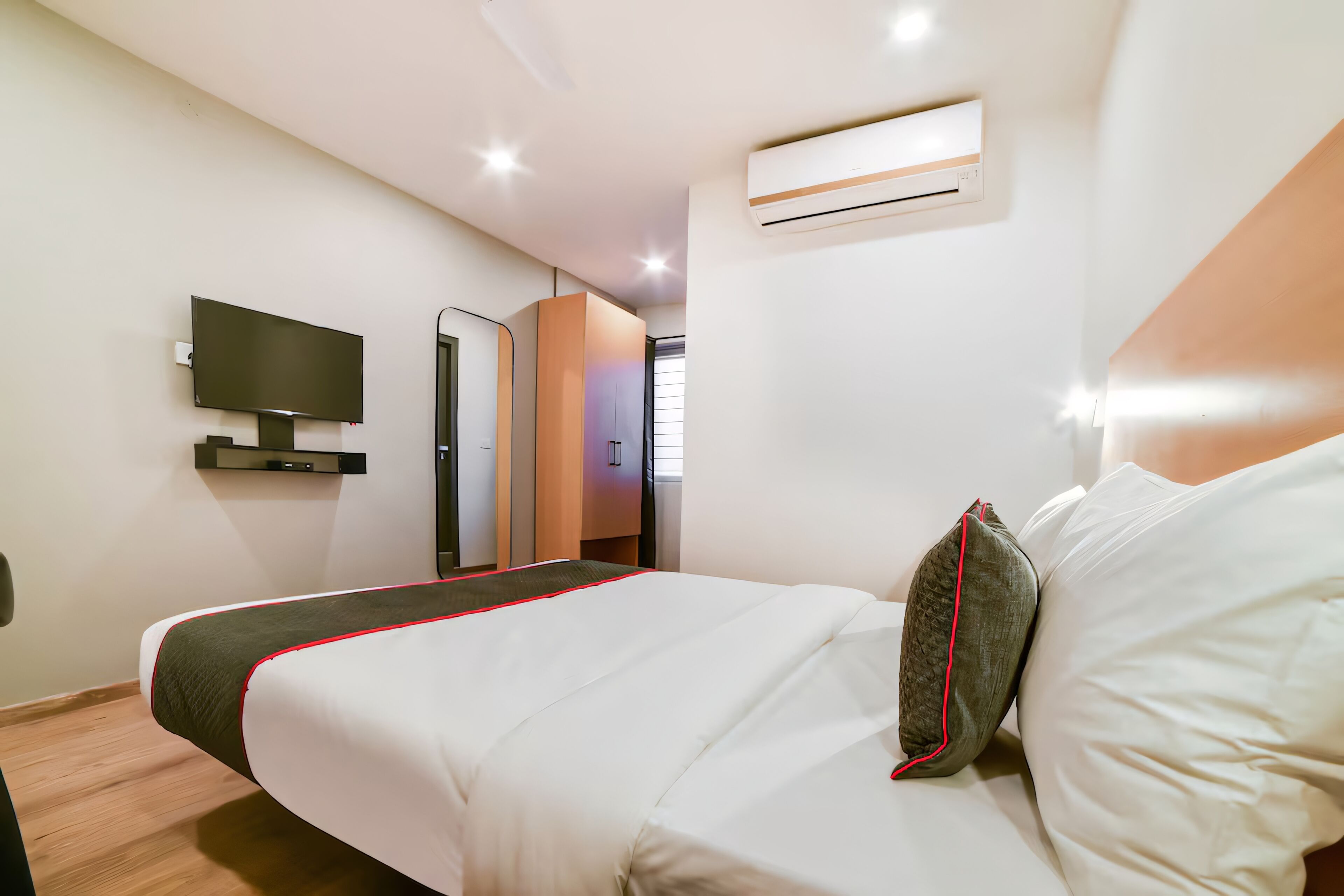 A modern hotel room with a large bed, a flat-screen TV mounted on the wall, and an air conditioning unit above the bed.