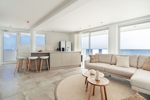 Un espacio habitable moderno con cocina, comedor y sofá frente a un ventanal con vista al mar.