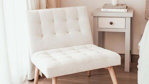 A white tufted chair with wooden legs in a room with light wooden flooring, a white bedside table with a drawer, and a black wall-mounted lamp.