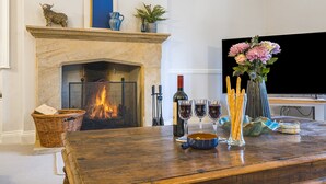 Een woonkamer met een open haard, een televisie en een houten tafel met wijnglazen en een bloevas.