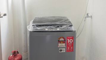 Un refrigerador compacto gris, motorizado, con una etiqueta de garantía de 10 años, un extintor y una tubería blanca sobre un pared blanco.