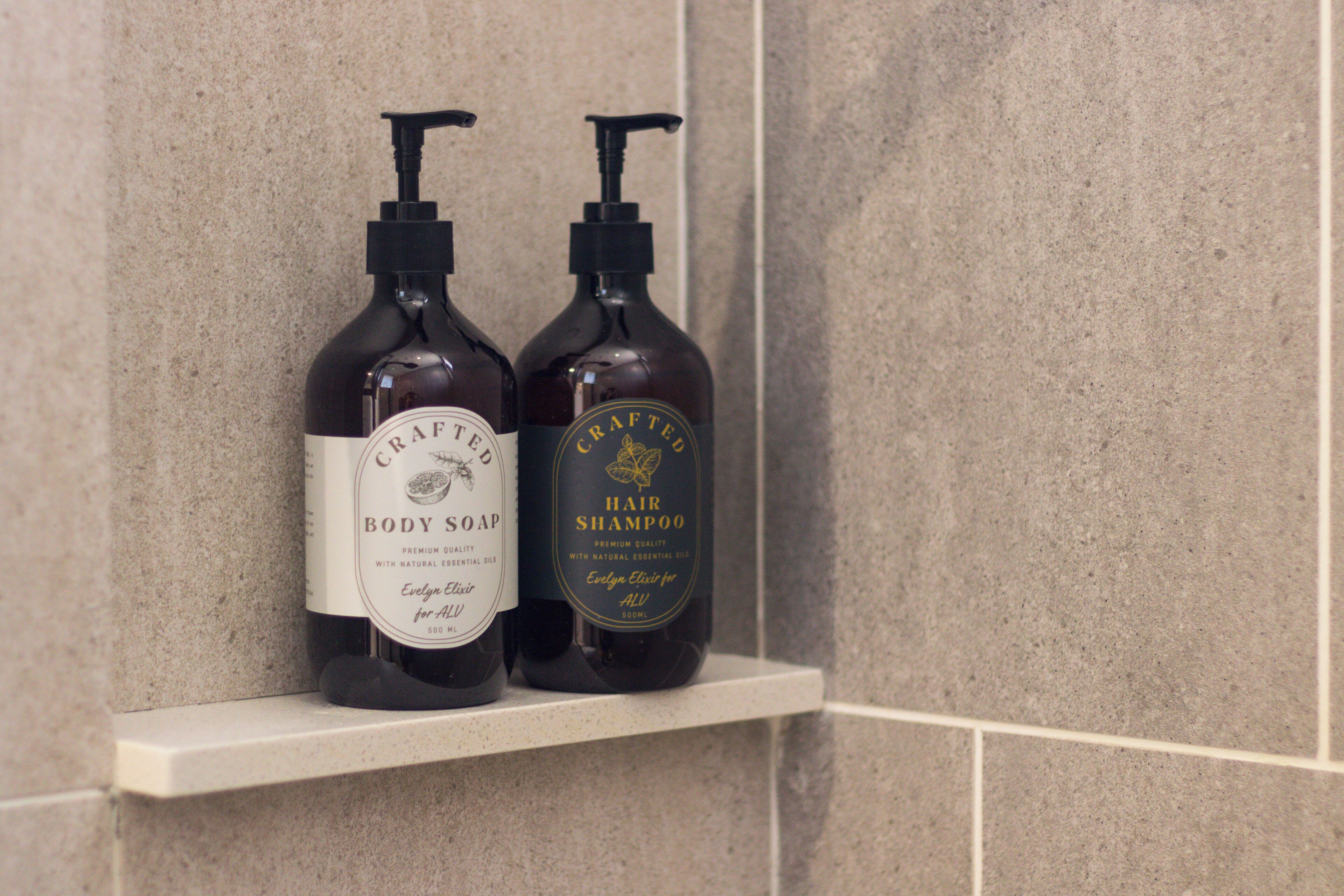Kaksi pulloa vartalosaippuaa ja hiusten shampoota valkoisella hyllyllä, taustalla laatoitettu seinä.