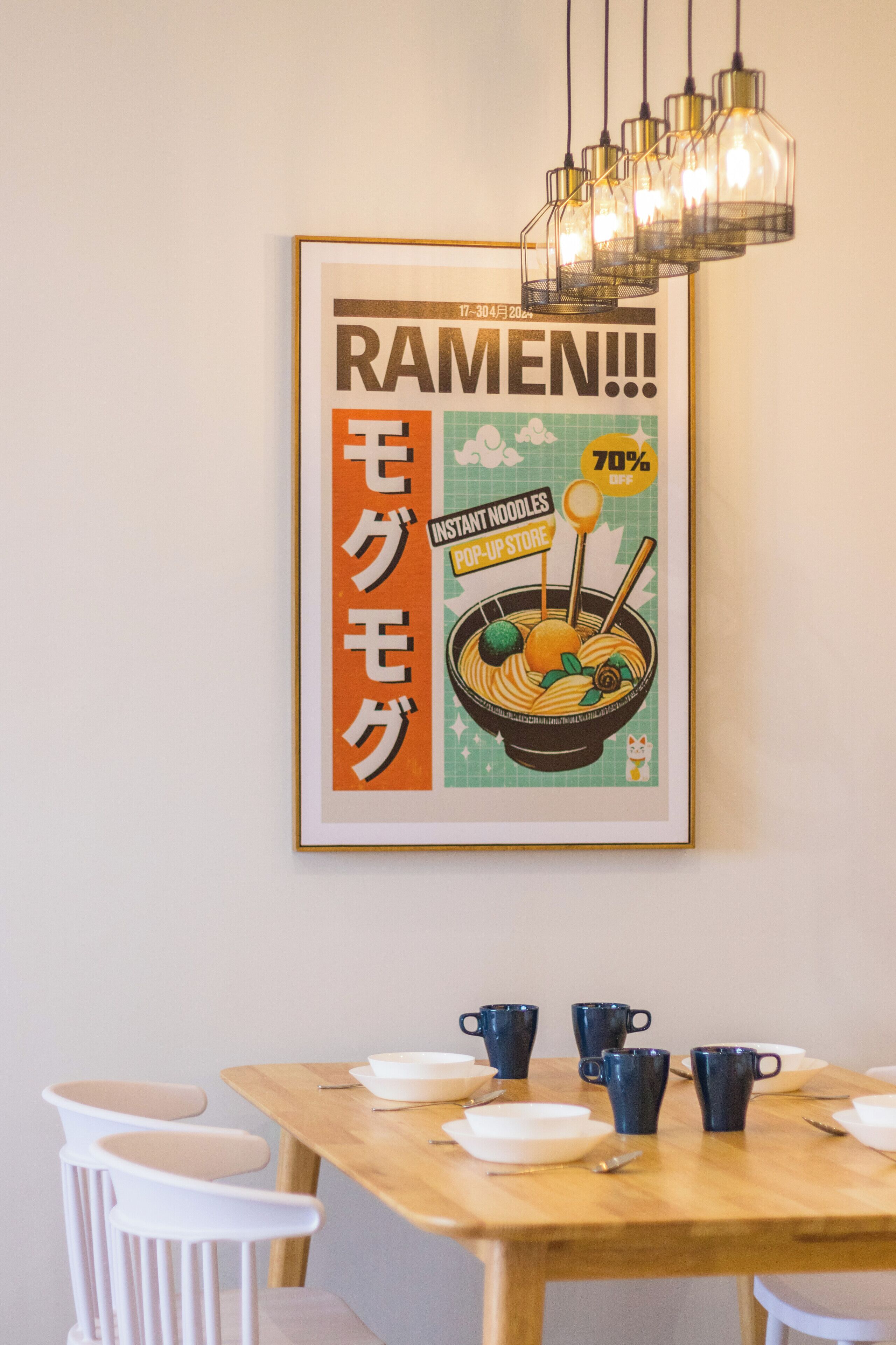 A dining area with a table set for four, a framed poster on the wall, and a hanging light fixture above.