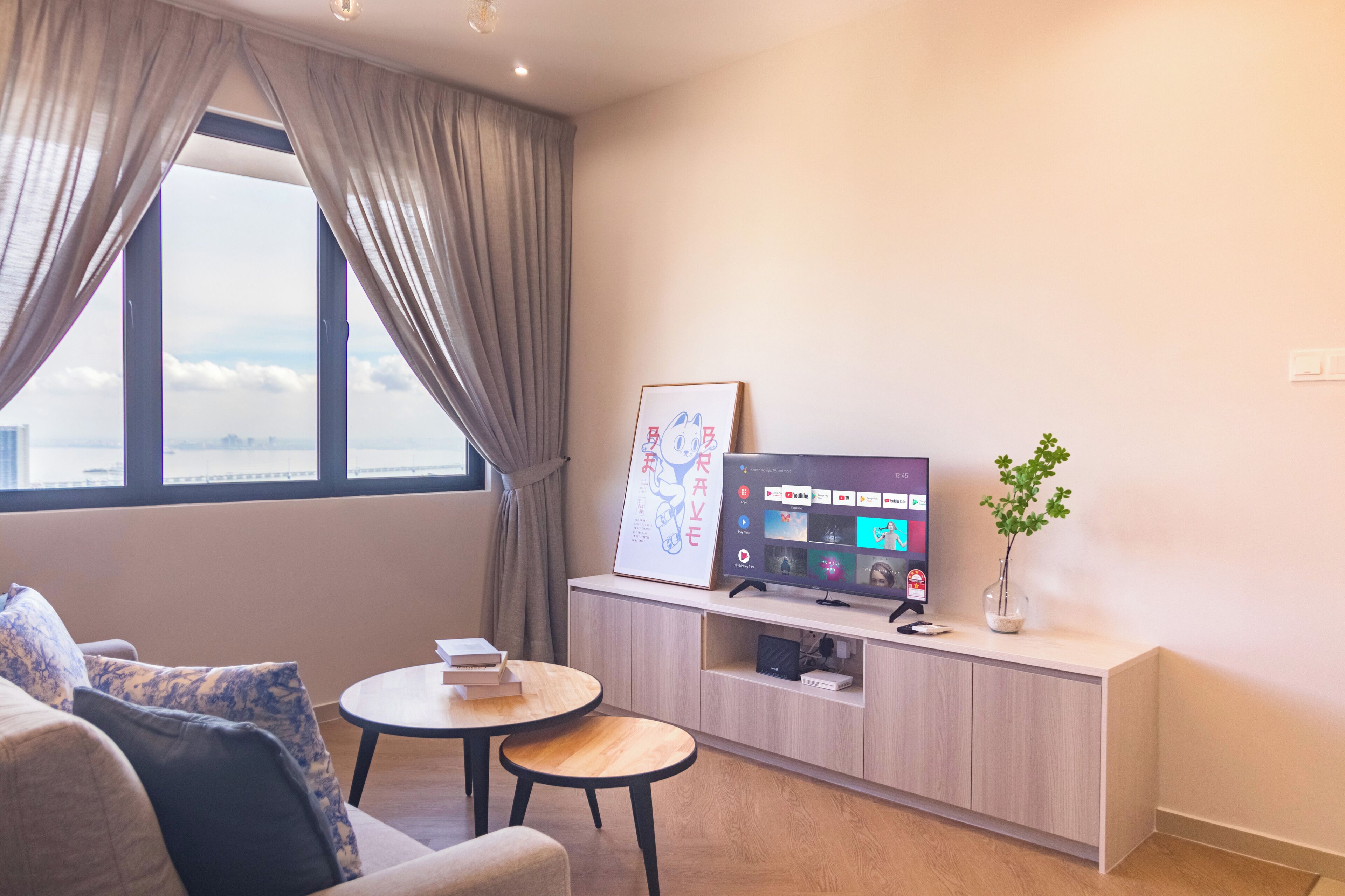 A modern living room with a sofa, two small tables, a mounted TV, and a large window with curtains.