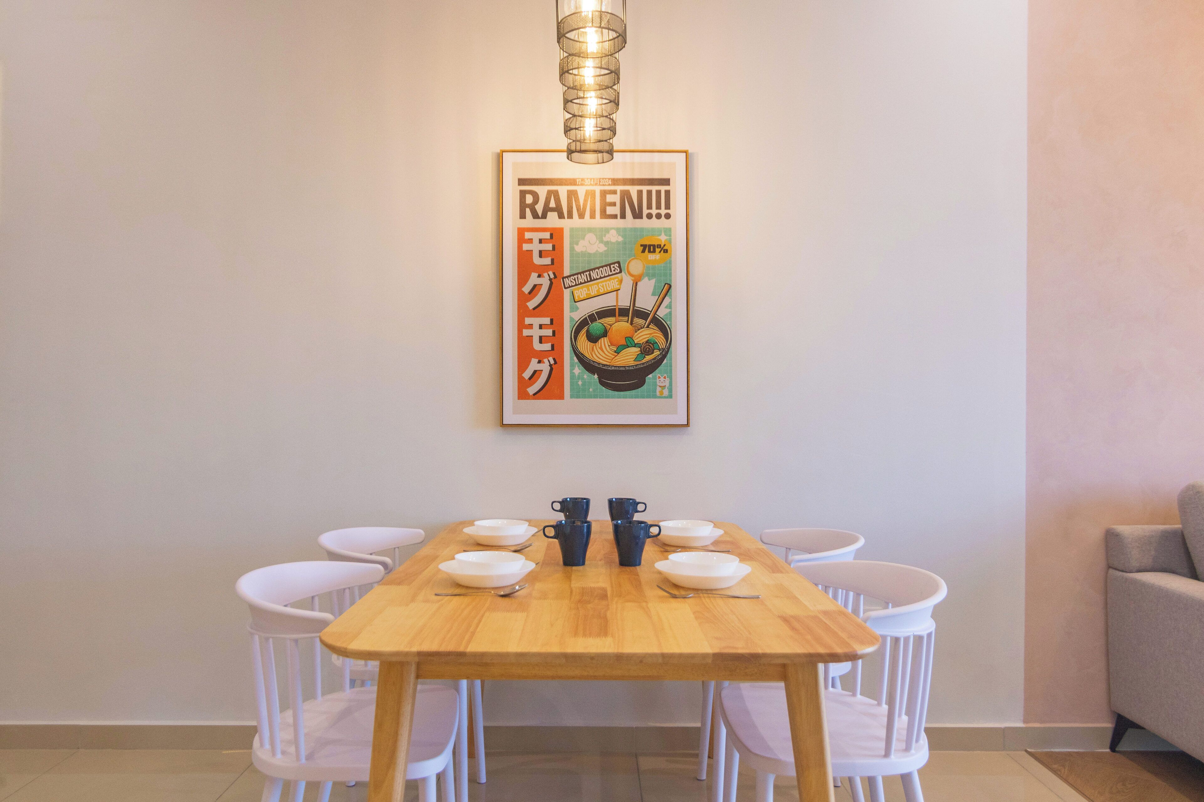 A dining area with a wooden table and white chairs, a framed poster on the wall, and a hanging light fixture.