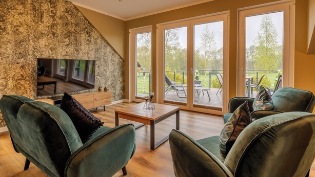A living room with a stone wall, a flat-screen TV, green upholstered chairs, a wooden coffee table, and large sliding glass doors leading to an outdoor patio. - Hünzinger Dorfschmiede - Holiday apartment Zaunkönig (Walsrode)