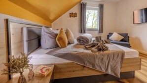 A modern bedroom with a wooden headboard, a large bed with multiple pillows, a bedside table with a vase and book, and a window with curtains. - Hünzinger Dorfschmiede - Holiday apartment Zaunkönig (Walsrode)
