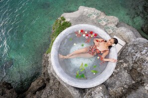 A person relaxing in a stone bathtub filled with water and flowers, surrounded by clear water and rocky terrain. - Casa Verde Cliff Resort and Spa (San Francisco)