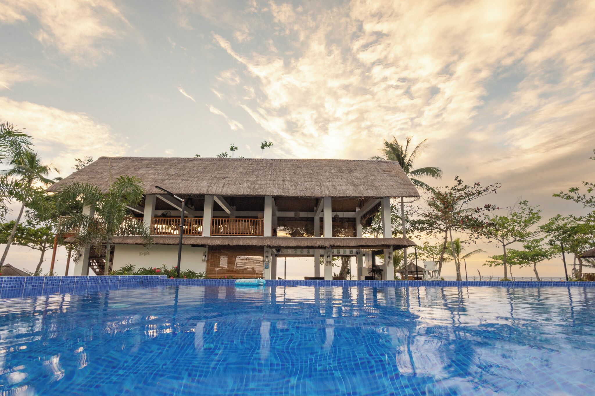 A resort with a large swimming pool, a thatched-roof building, and palm trees.