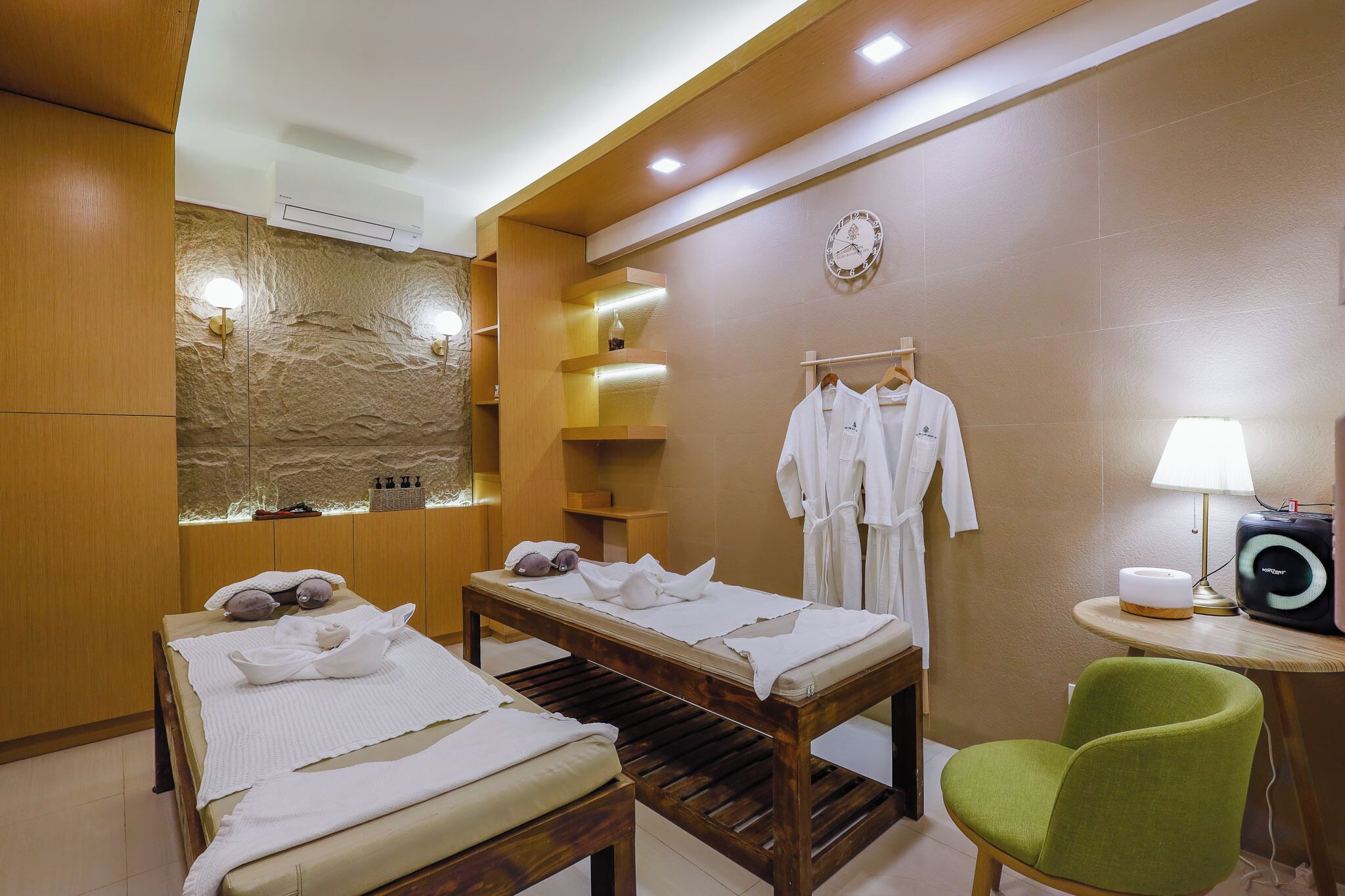 A spa room with two massage tables, a green chair, a wall-mounted clock, and a small table with a lamp and a speaker.