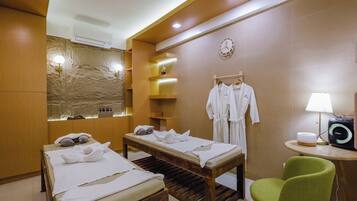 A spa room with two massage tables, a green chair, a wall-mounted clock, and a small table with a lamp and a speaker.