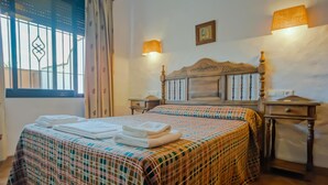 Una cama bien hecha con colcha escocesa y toallas dobladas, dos lámparas de noche, un cabecero de madera y una ventana con cortinas.