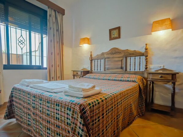 A neatly made bed with a plaid bedspread and folded towels, two bedside lamps, a wooden headboard, and a window with curtains.