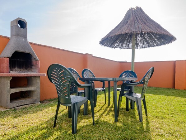 Un patio al aire libre con una parrilla de piedra, una mesa con sillas y un parasol de paja.