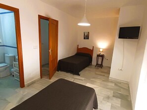 Un dormitorio con cama, mesita de noche con lámpara, televisor montado en la pared y baño visible a través de una puerta abierta.