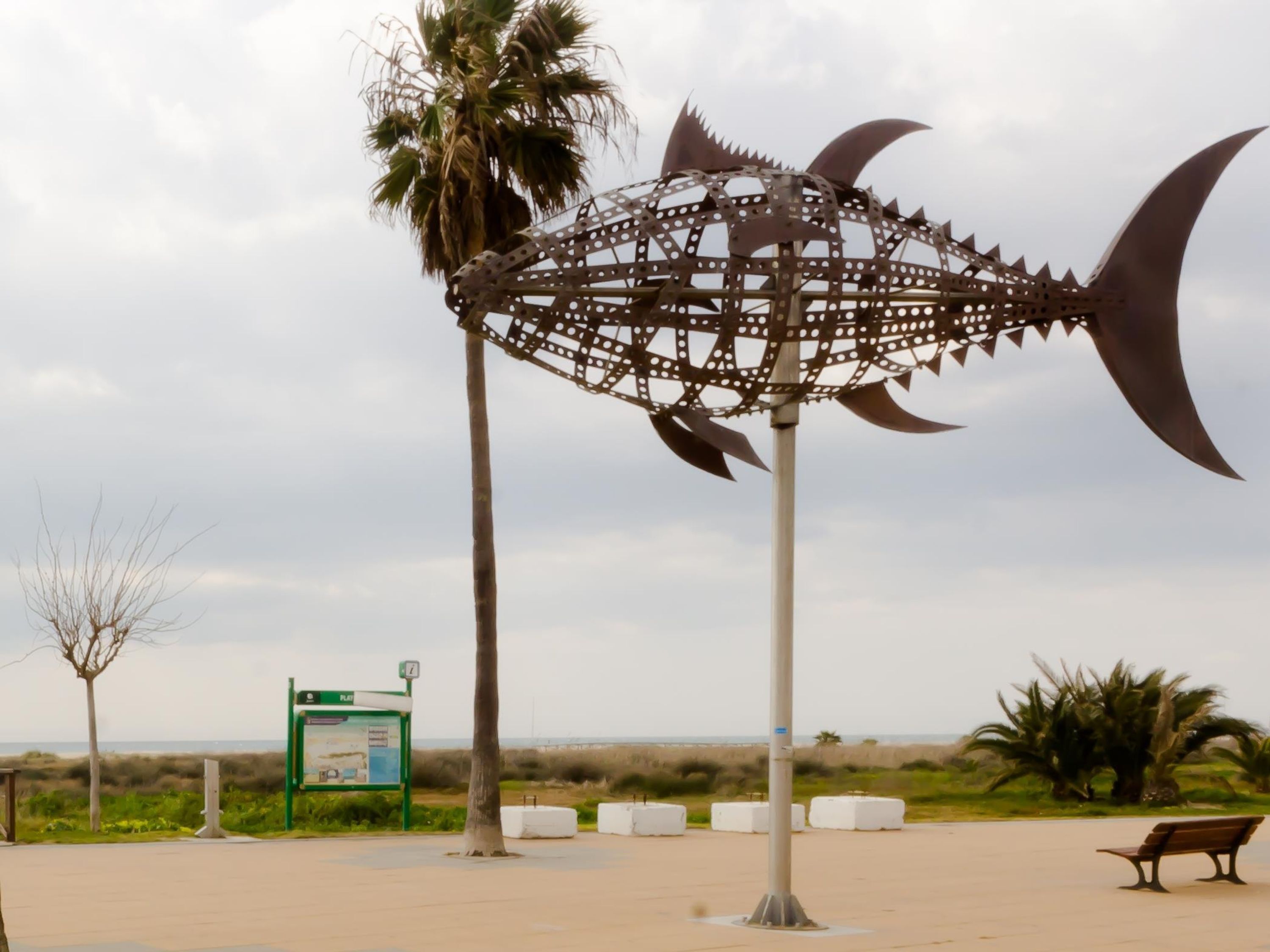 Eine Metallskulptur, die einen Fisch darstellt, mit einer runden Öffnung, einer Bank und einem Hinweisschild in einer Parkanlage.
