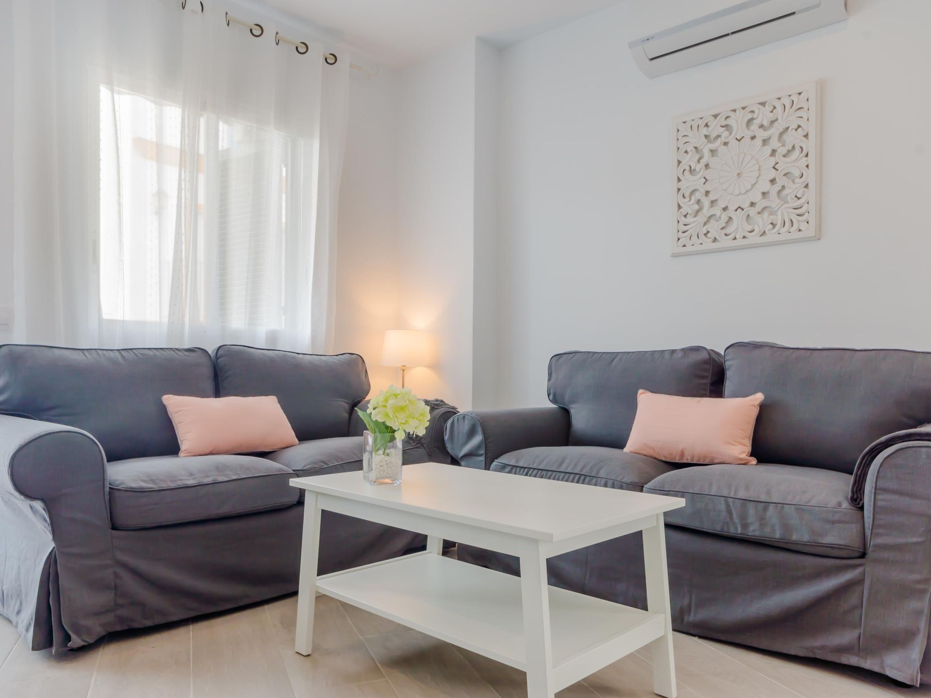 Ein modernes Wohnzimmer mit zwei dunkelgrauen Sofas, jedes mit einem hellrosa Kissen, einem weißen Couchtisch und einer Vase mit Blumen.