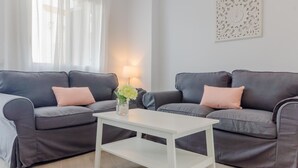 Una sala de estar moderna con dos sofás grises oscuros, cada uno con un cojín rosa claro, una mesa de centro blanca y un jarrón con flores.