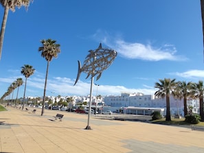 Un paseo marítimo con palmeras, una escultura de metal y un edificio con la leyenda "NOSTAL" al fondo.