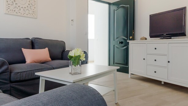 Una sala de estar moderna con un sofá gris oscuro, una mesa de centro blanca, un otoman gris, un mueble blanco con un televisor de pantalla plana y un panel decorativo blanco en la pared.