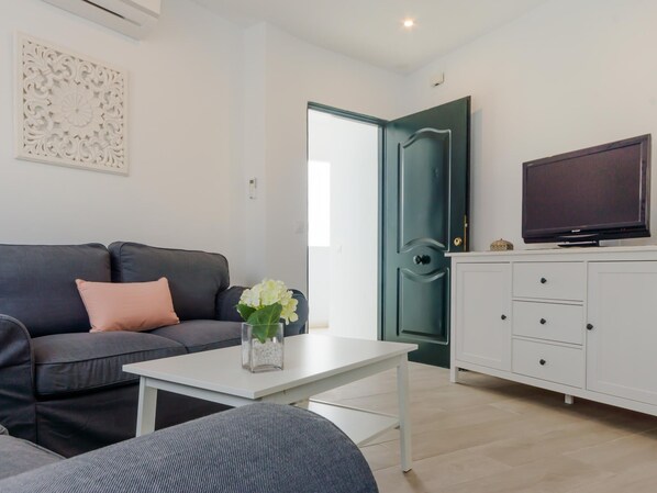 Ein modernes Wohnzimmer mit einem dunkelgrauen Sofa, einem weißen Couchtisch, einem grauen Hocker, einer weißen Kommode mit Flachbildfernseher und einer dekorativen weißen Wandverkleidung.