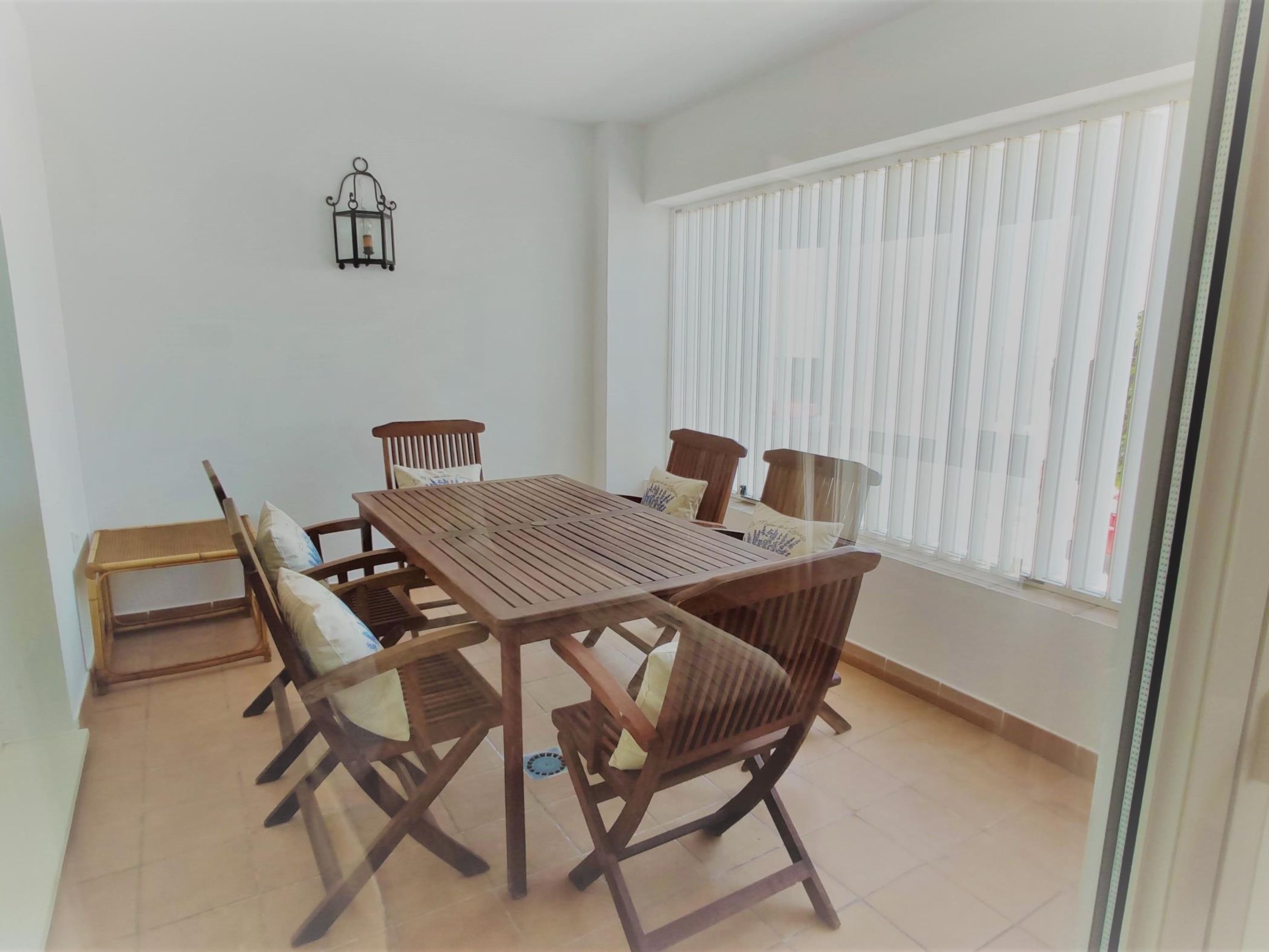 Un comedor con una mesa de madera y sillas, una mesita auxiliar y una ventana con persianas verticales.